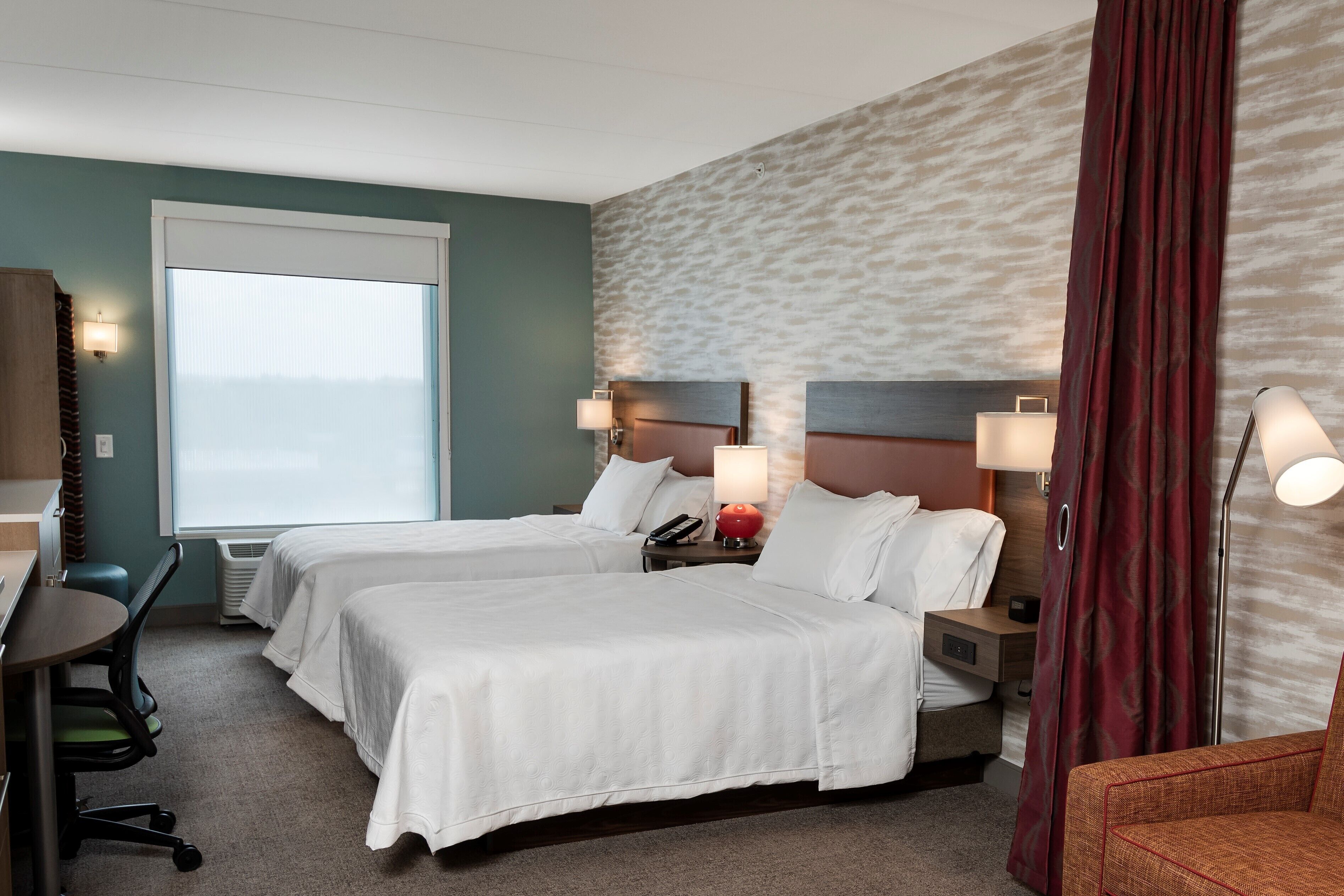 Ein Hotelzimmer mit zwei Betten, einem Schreibtisch und einem Stuhl. Zwei Nachttische mit Lampen und einem Telefon sind vorhanden.