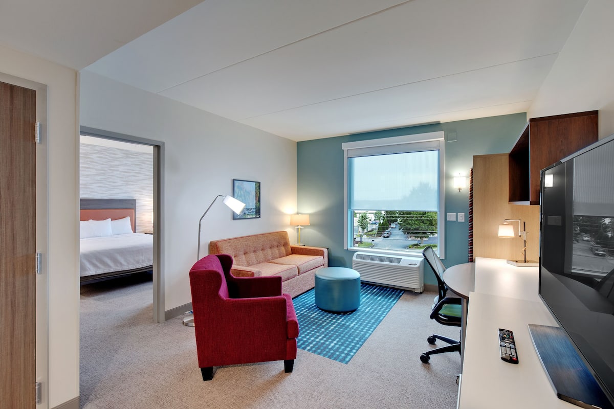 Une chambre d’hôtel avec un lit, un fauteuil rouge, un canapé, un bureau avec une télévision et une grande fenêtre donnant sur l’extérieur.