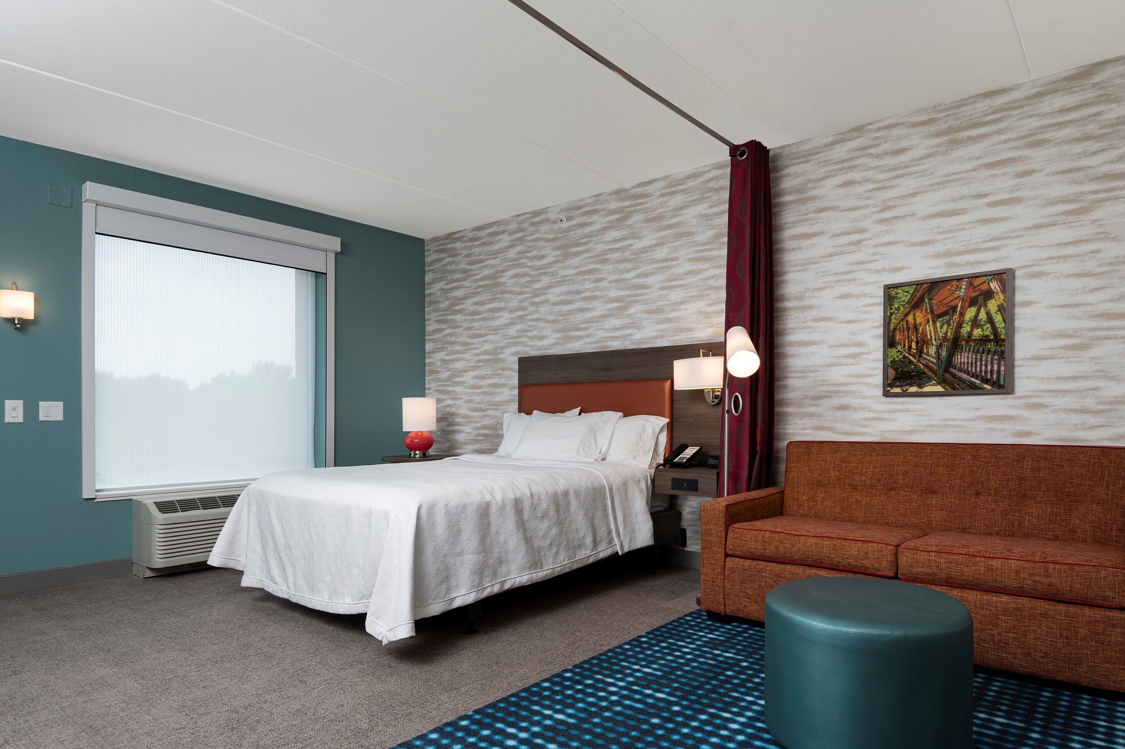 A hotel room with a bed, a sofa, a nightstand with a lamp, a wall-mounted phone, and a window with blinds.