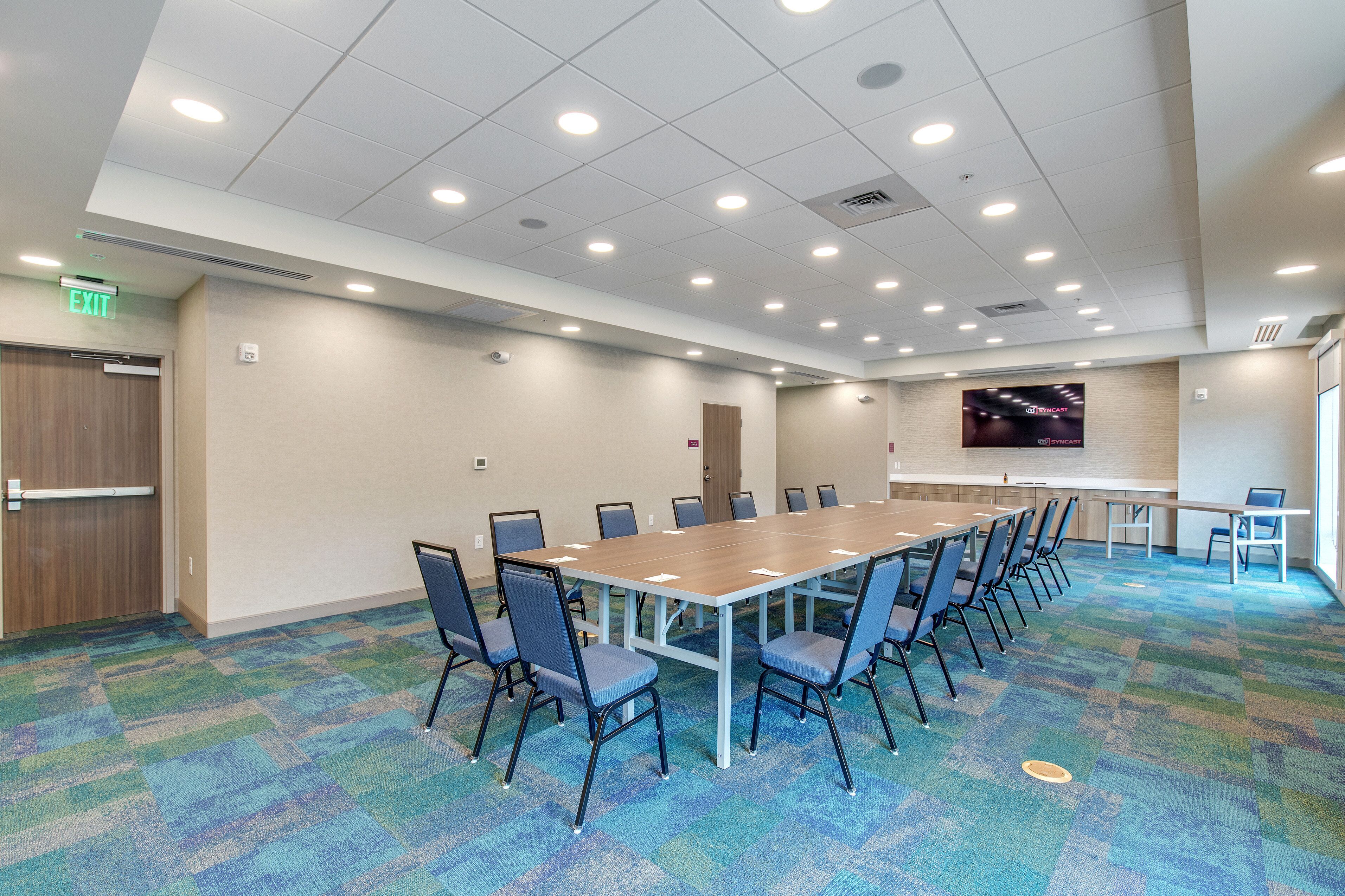 A conference room with a long table, chairs, carpeted floor, and a TV screen.