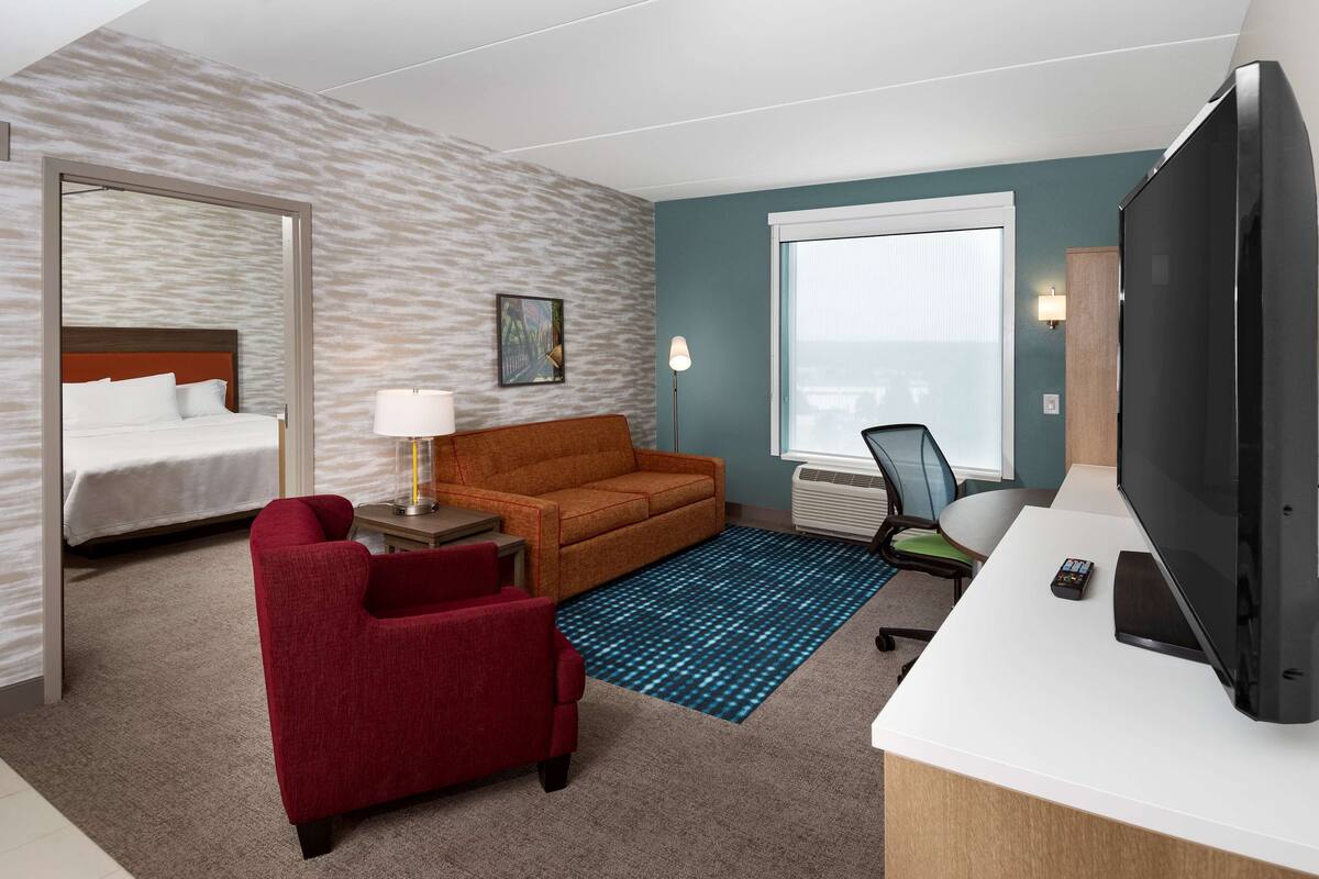 Une chambre d’hôtel avec un lit, un fauteuil rouge, un canapé, un bureau avec une télévision et une fenêtre.