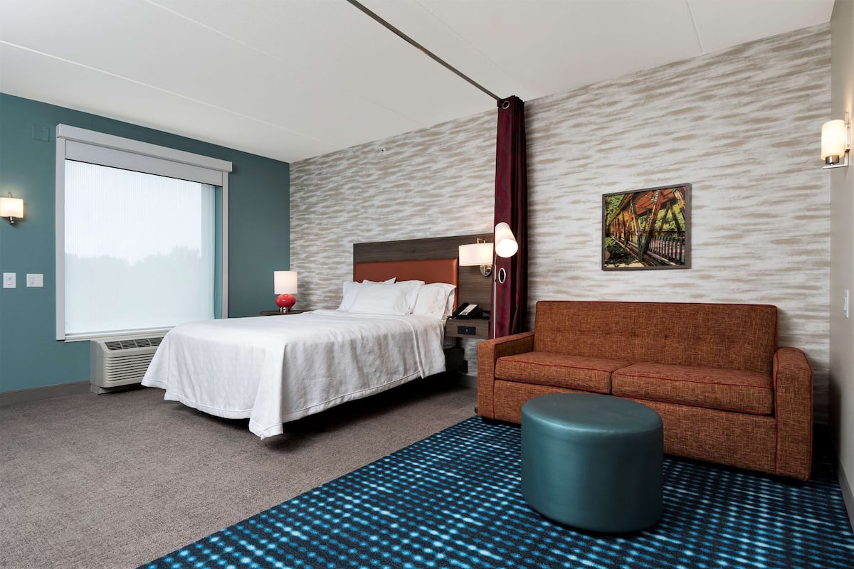 Une chambre d’hôtel avec un lit, un canapé, un pouf rond, un téléviseur fixé au mur et un tableau.