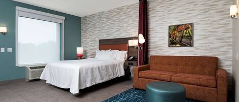 Une chambre d’hôtel avec un lit, un canapé, un pouf rond, un téléviseur fixé au mur et un tableau.