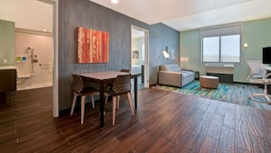 A modern hotel room with a dining area, a sofa, a TV, and a large window.