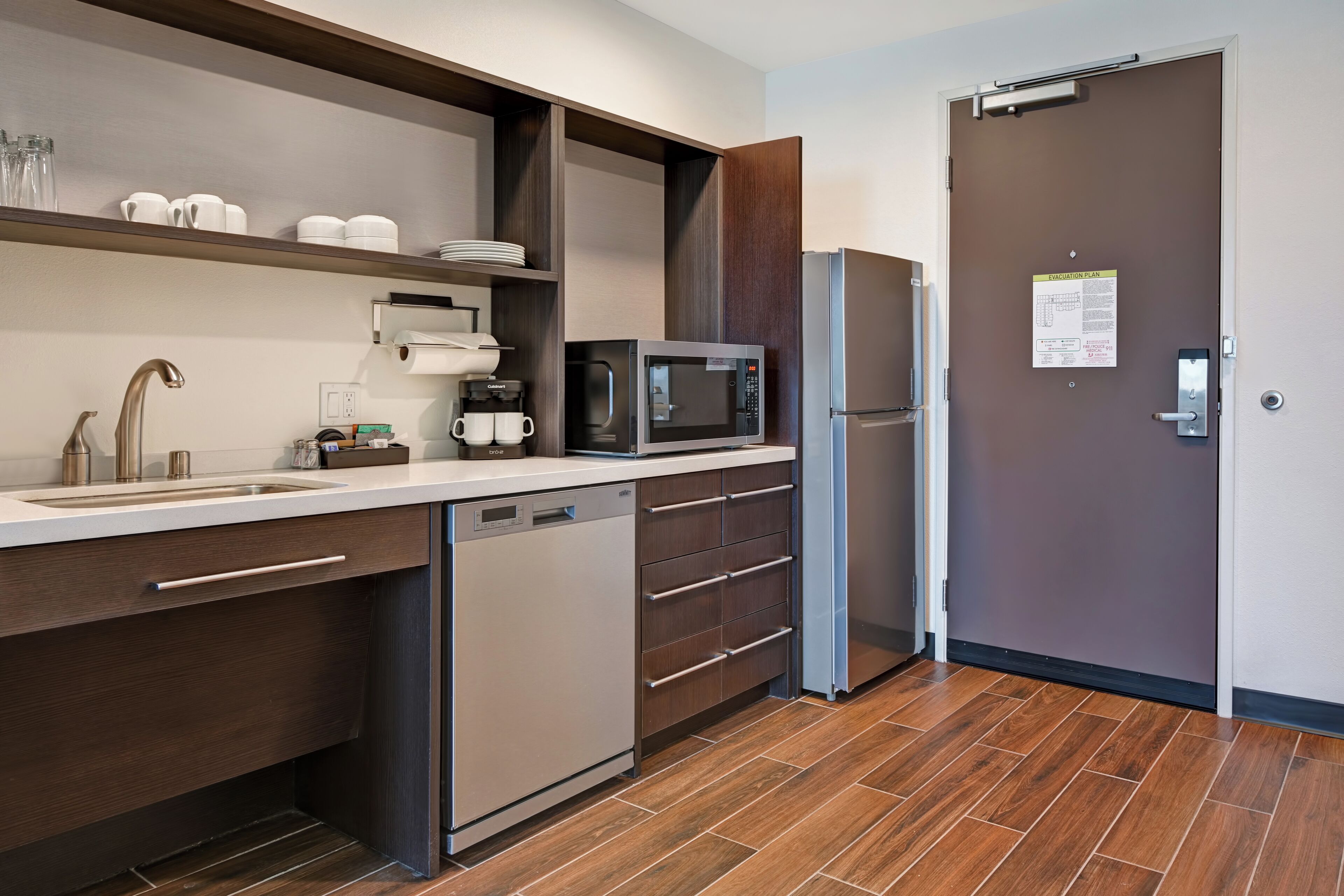 A modern kitchen with a dishwasher, microwave, and refrigerator. There is a sink with a faucet, and a cabinet above the sink.