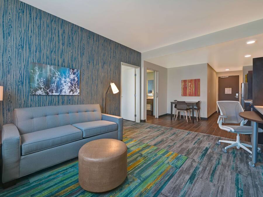 A modern hotel room with a blue sofa, a round ottoman, a desk with a chair, and a dining area.