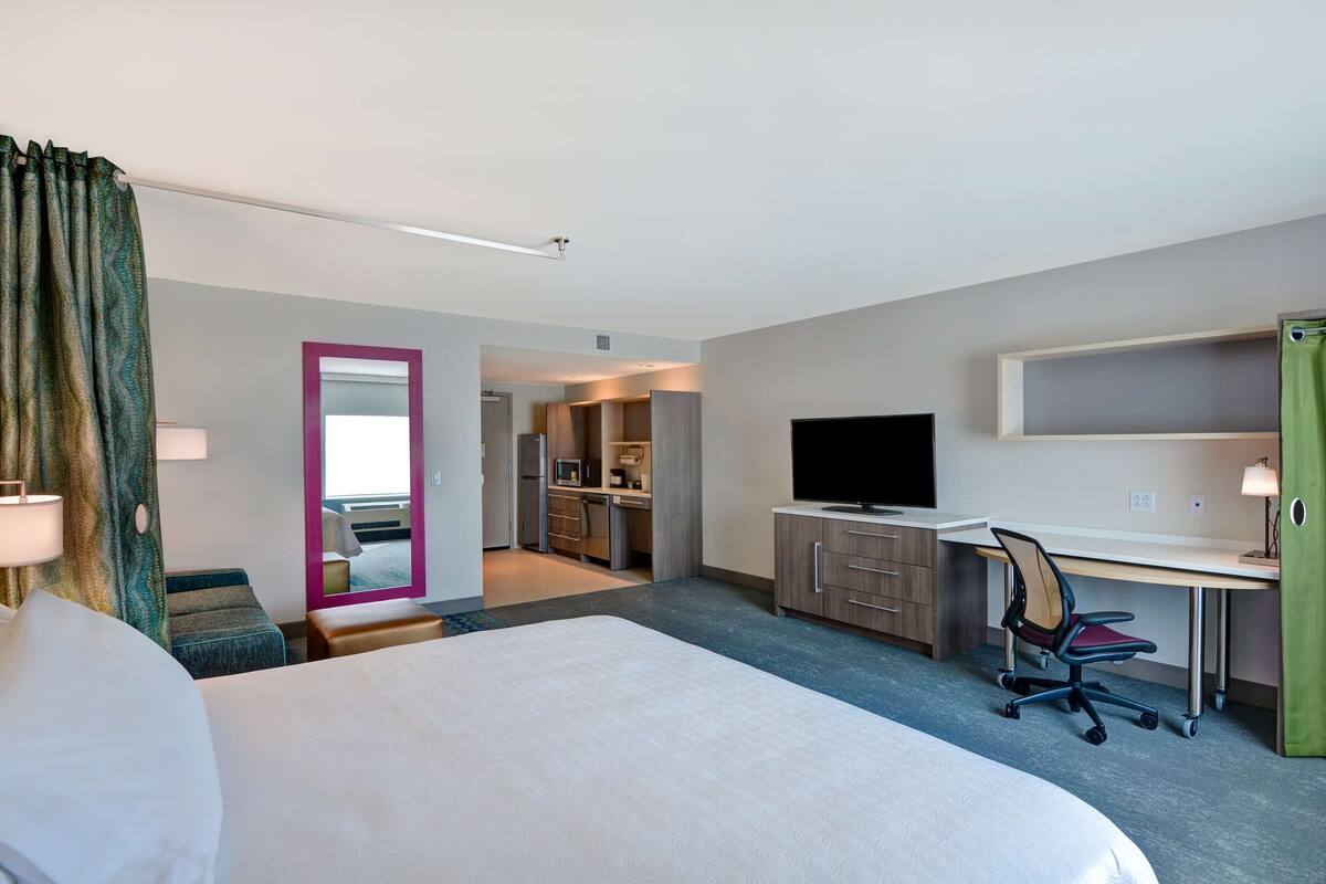 Ein Hotelzimmer mit Bett, Schreibtisch, Stuhl, Fernseher und einer kleinen Küche im Hintergrund.