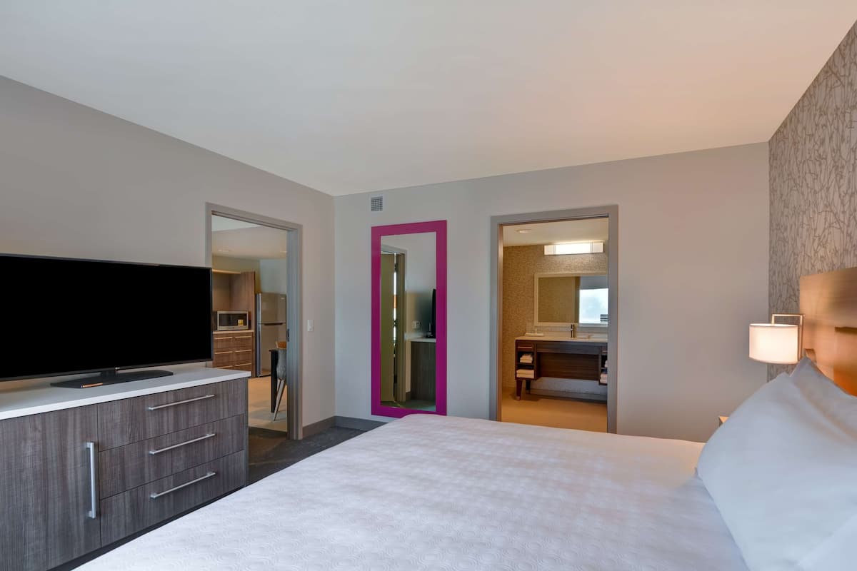 Ein Hotelzimmer mit einem großen Bett, einem Fernseher auf einer Kommode, einem Badezimmer mit Spiegel und einer Tür zu einem weiteren Raum.