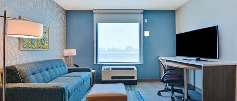 A hotel room with a blue sofa, a small table with a lamp, a desk with a computer and TV, and a large window with a view.