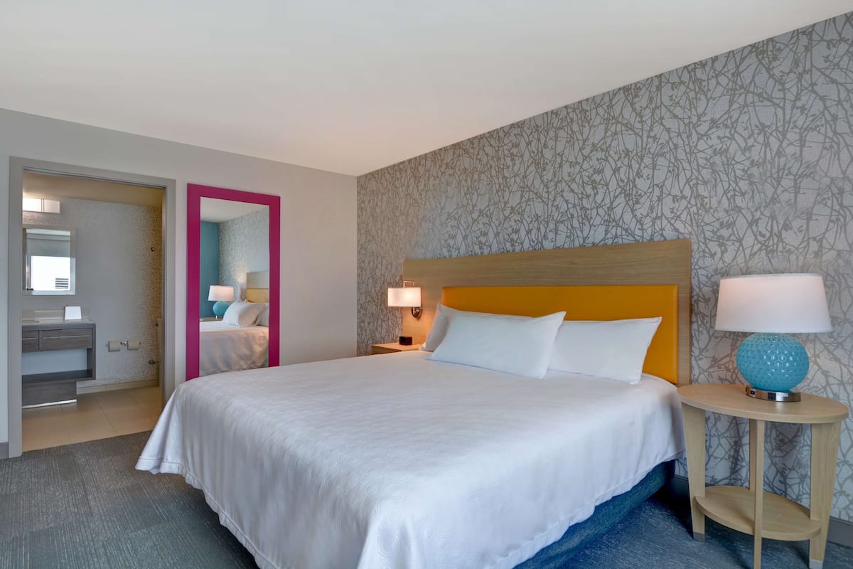 Ein Hotelzimmer mit einem großen Bett, einem hölzernen Kopfteil, einem Nachttisch mit Lampe und einem durch eine offene Tür einsehbaren Badezimmer.