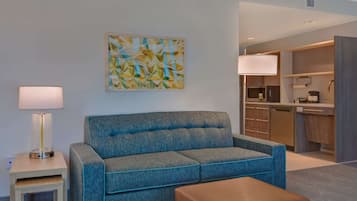 A modern hotel room with a blue sofa, a small table, a lamp, and a kitchen area in the background.
