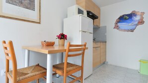 Een kleine kitchenette met een witte koelkast, een magnetron en houten kastjes. Er staat een tafel met twee rieten stoelen bij, en een vaas met bloemen. Een schilderij hangt aan de muur boven de tafel.