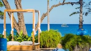 Un área junto a la piscina con una silla mecedora de madera, abundante follaje y vista al mar con un barco a lo lejos.