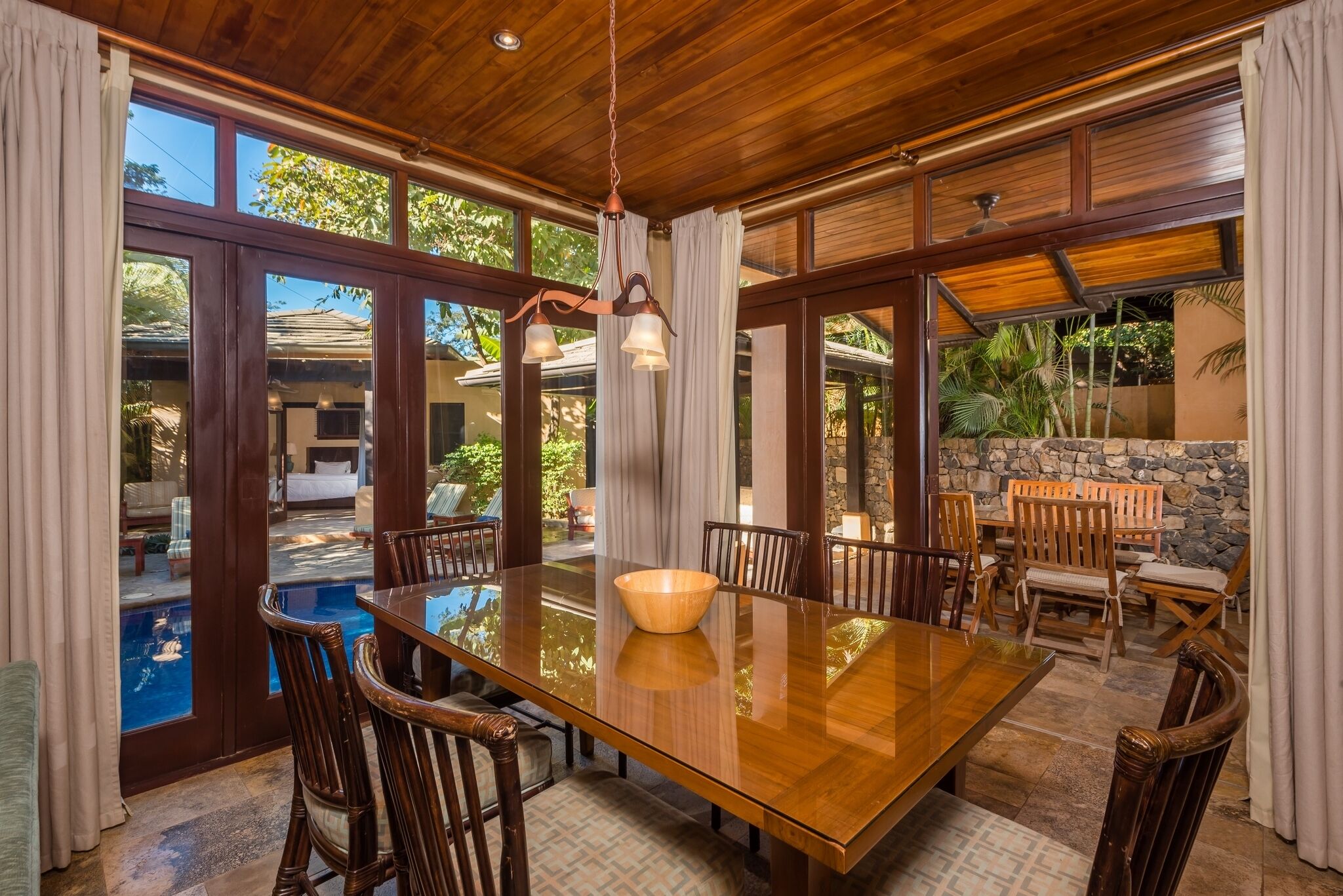 A spacious dining area with a large wooden table and chairs, a hanging light fixture, and a view of a swimming pool and outdoor seating area.
