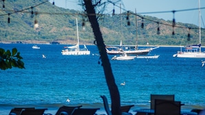 Vista al mar con barcos, un comedor con sillas y montañas al fondo.