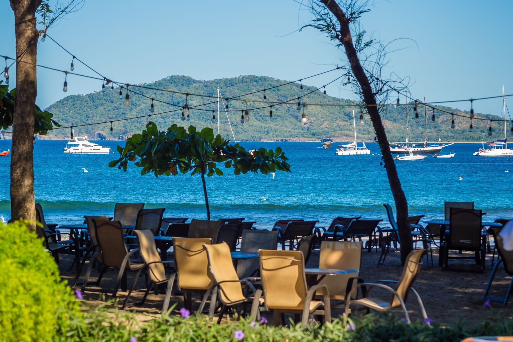 Outdoor seating with tables and chairs, overlooking a body of water with boats and a hilly landscape in the background.