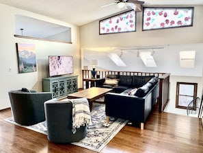 A living room with a sofa, ottoman, coffee table, and a TV. There are two windows with curtains, a ceiling fan, and a rug on the floor. - Family Escape with Hot Tub, Arcade Room & Wildlife Views Sleeps 16, Pets OK (Colorado Springs)