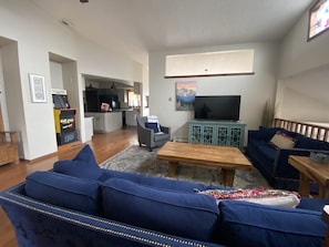 A living room with a blue sofa, a wooden coffee table, and a television. There is a kitchen in the background and a Pac-Man arcade machine. - Family Escape with Hot Tub, Arcade Room & Wildlife Views Sleeps 16, Pets OK (Colorado Springs)