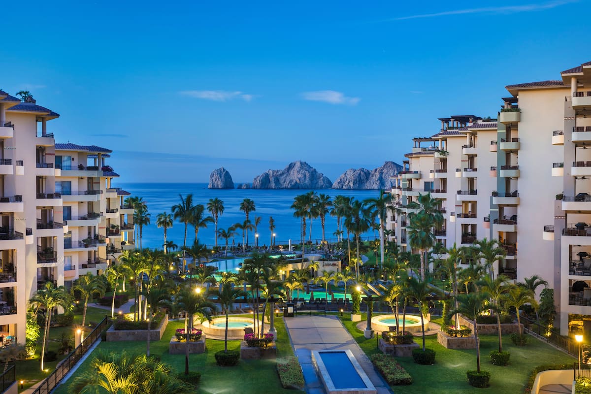 Un resort con varias edificaciones, piscina, palmeras y vista al océano.
