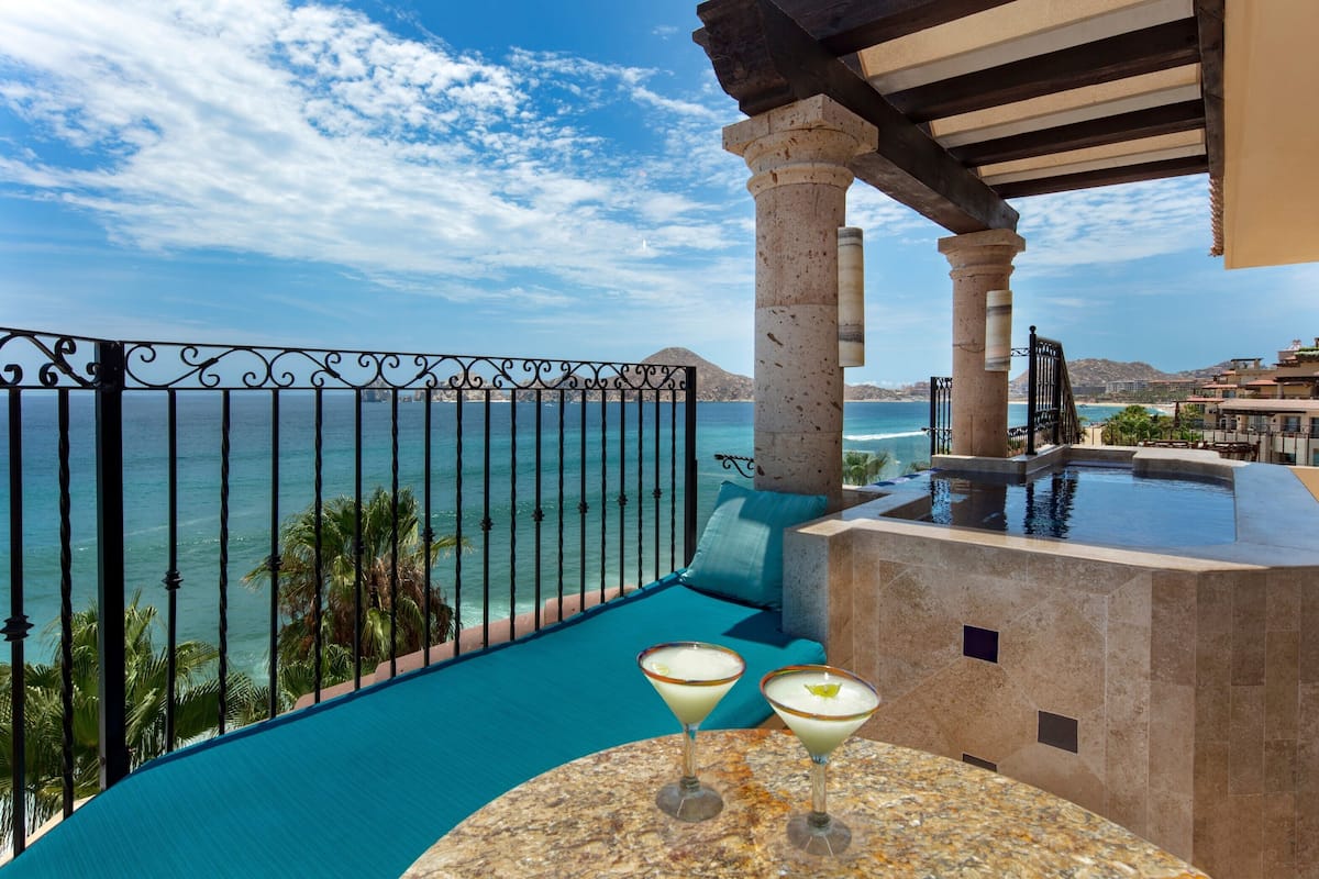 Un balcón con vista al mar, una piscina y dos bebidas sobre una mesa.