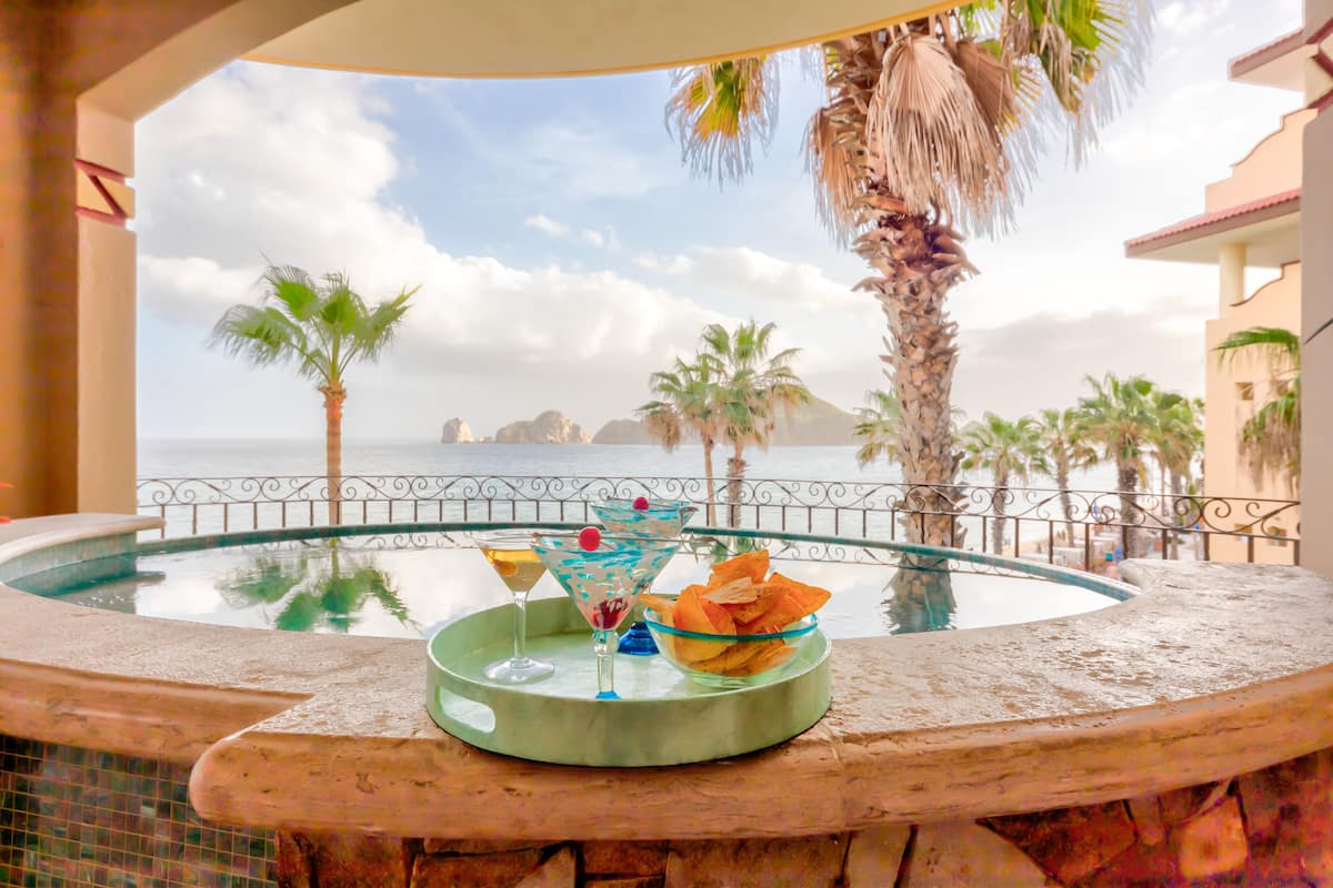 Zona de piscina con vista al mar, palmeras y una mesa de buffet con bebidas y aperitivos.