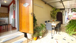 Un patio con puerta de madera, mobiliario exterior, plantas en macetas y suelo de baldosas.