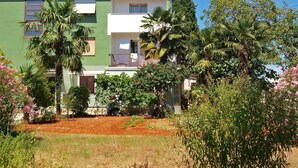 Un edificio de dos pisos con fachada verde, un balcón con barandilla y un pequeño jardín con palmeras y plantas en flor.
