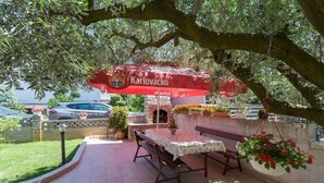 An outdoor seating area with a table and benches under a red umbrella with 'Karlovacko' branding, surrounded by greenery and a brick oven.