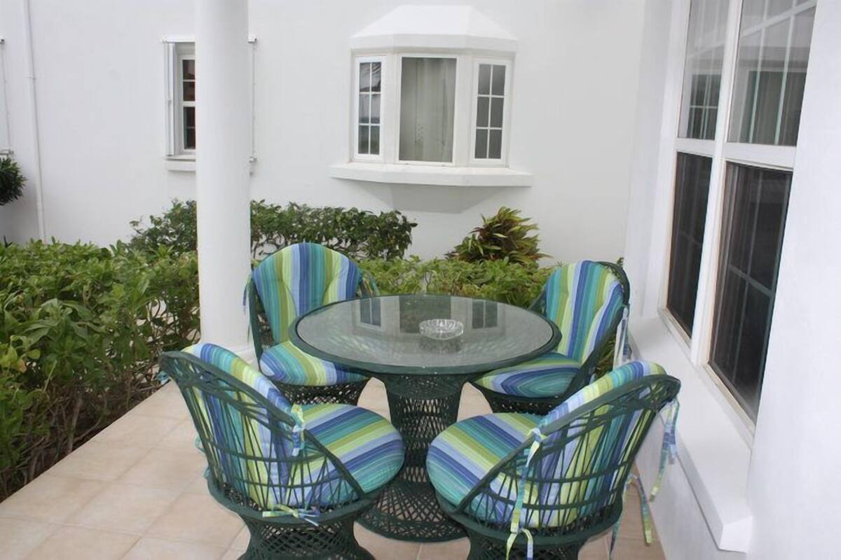 Une table ronde en verre, entourée de quatre chaises dotées de coussins à rayures, est placée sur une terrasse aux murs blancs et agrémentée d’une grande baie vitrée.