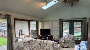 A spacious living room with a large TV, floral sofas, and a view of a swimming pool. - The Lake Tapps Bungalow (Lake Tapps)
