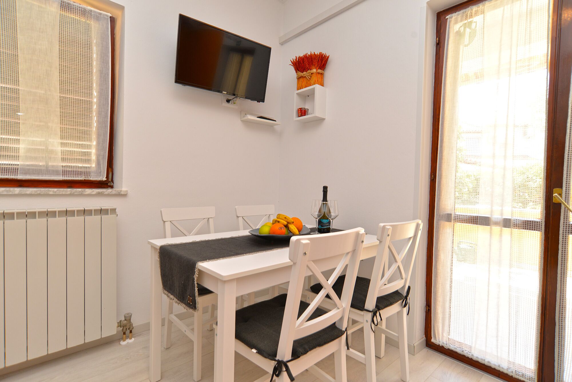 Un comedor con una mesa, sillas, un televisor montado en la pared y un ventanal con cortinas.