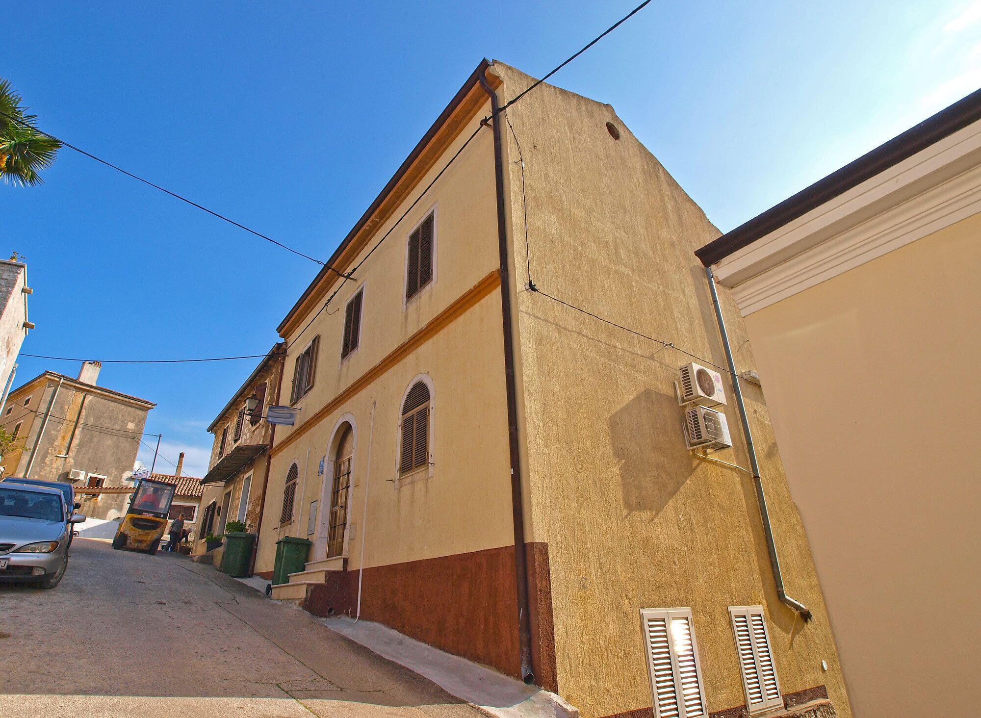 A narrow street with buildings on either side, featuring a mix of architectural styles, including a building with a beige facade and a brown base.
