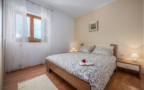 A bedroom with a bed, bedside tables, a window with sheer curtains, a bedside lamp, and a framed picture on the wall.