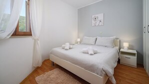 Une chambre à coucher comprenant un lit, des tables de chevet, une commode, une fenêtre avec des rideaux légers, une œuvre d’art accrochée au mur et un plancher en bois.
