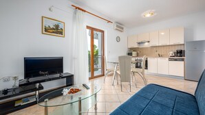Un soggiorno moderno con TV, tavolino in vetro e divano blu. Si intravede sullo sfondo una cucina con mobili e elettrodomestici bianchi.