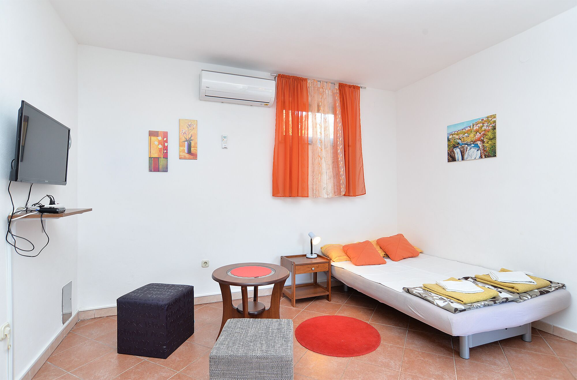 Une chambre de taille modeste, équipée d’un lit, d’un bureau, d’une chaise, d’un climatiseur et d’un téléviseur.