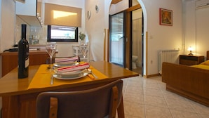 A dining area with a wooden table, chairs, and a bottle of wine. There is a window with blinds, a door leading to a bathroom, and a radiator on the wall.