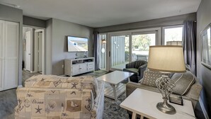 A living room with a television, sofa, and coffee table. There is a door leading to another room and a window with curtains.