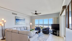 A living room with a sofa, two armchairs, a coffee table, a TV, and a large window with a beach view.