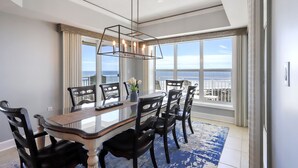 A dining room with a large table, black chairs, a chandelier, and a view of the ocean through large windows.