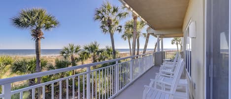 A balcony with white rocking chairs overlooking a beach with palm trees and the ocean.