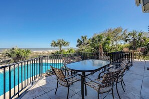 A poolside area with a table and chairs, a view of the ocean, and palm trees.