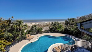 Piscina de borde curvo rodeada por una cerca metálica y con vistas a una playa con dunas y el océano.