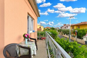 Un balcón con una mesa y sillas, un florero con flores y vistas a una calle con casas y follaje.