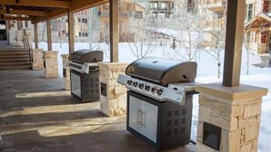 Deux barbecues d’extérieur sont installés sous une terrasse couverte, soutenue par des piliers en pierre, le tout se détachant sur un paysage enneigé avec des bâtiments visibles au loin.