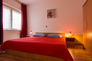 A bedroom with a red bedspread, two bedside lamps, a wooden wardrobe, and a window with red curtains.