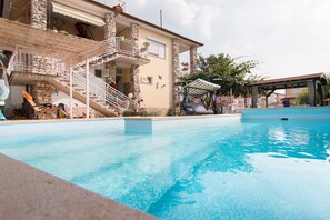 A swimming pool with a stone building in the background, a covered outdoor area, and a staircase leading to a balcony.