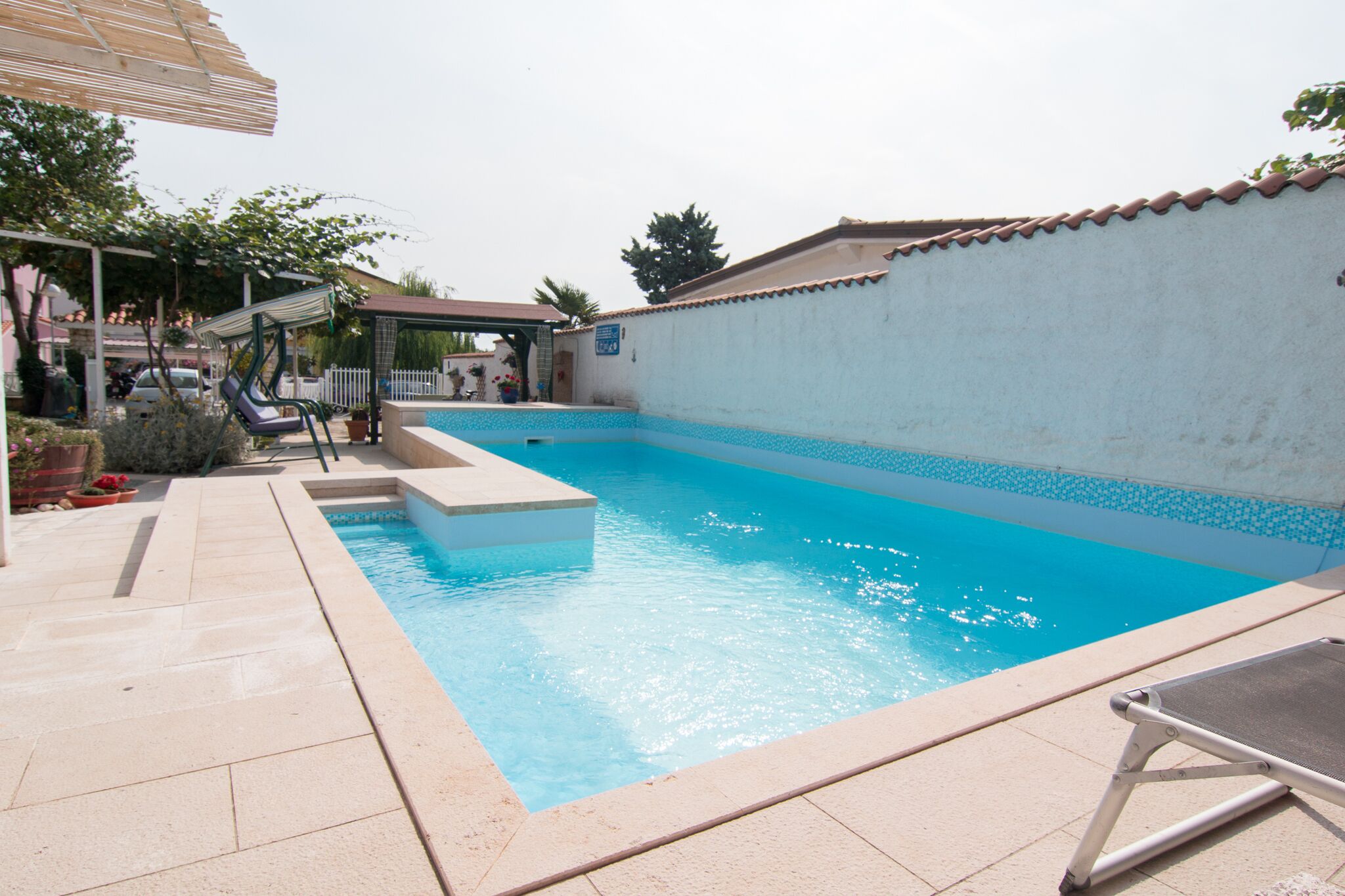 A swimming pool with a tiled deck, a covered seating area, and a white wall with a tiled roof.