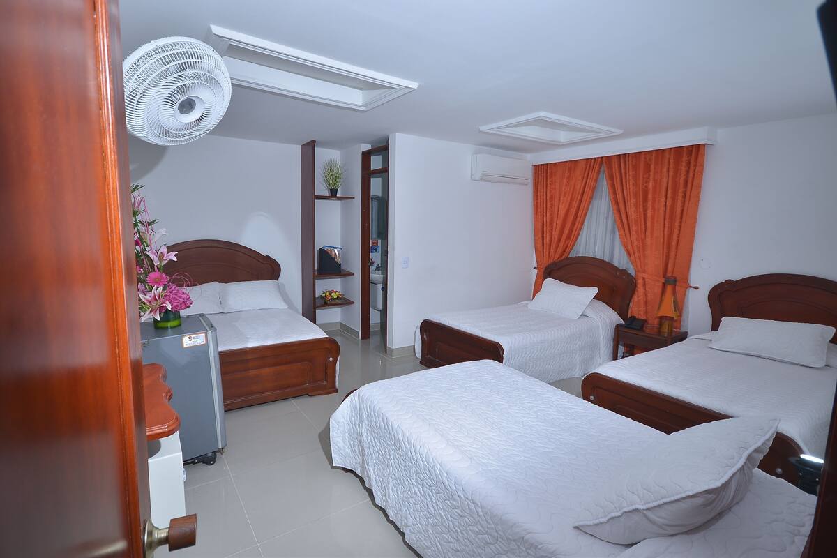 Hotelkamer met drie bedden, een houten garderobe, een klein bureau, een ventilator en een kamerplant.