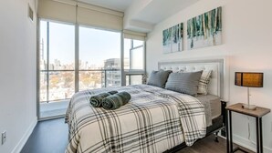 A bedroom with a large window, a bed with a plaid bedspread, a headboard, and a painting on the wall.